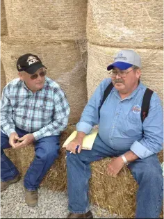 Two men sitting on hay bales in a barn, wearing caps and plaid shirts, talking casually.