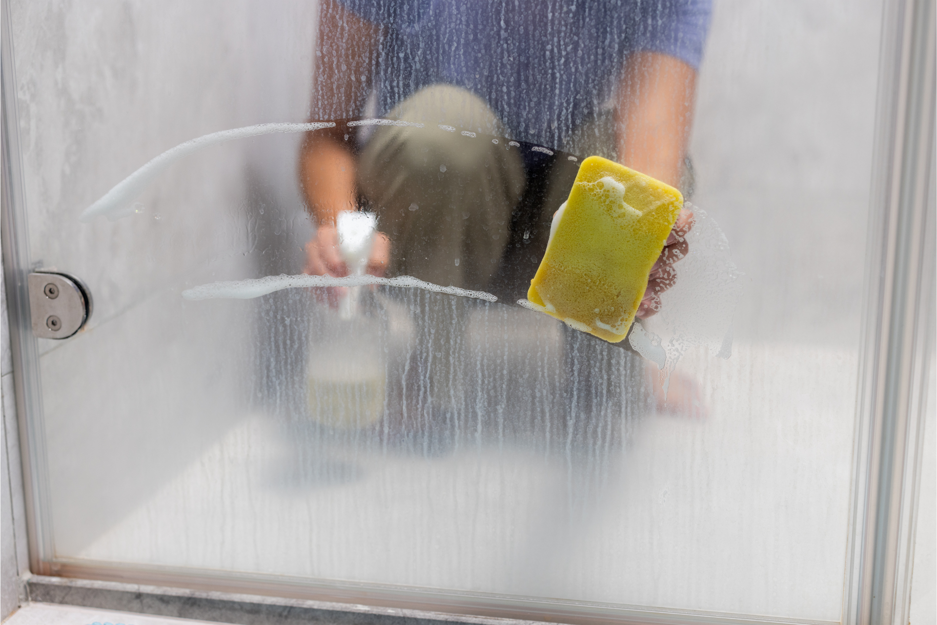 A man is cleaning a shower door with a sponge and spray.