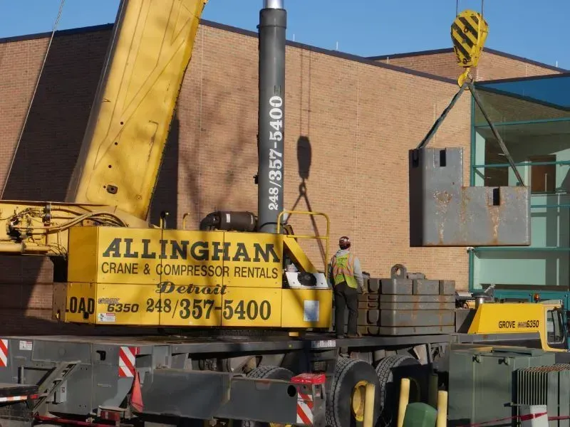Yellow crane lifting metal blocks near a building. Man in safety vest nearby.