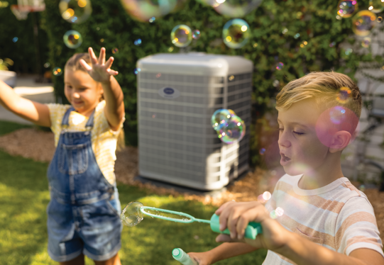 Two children blowing bubbles in a sunny backyard, with a window air conditioner in the background.