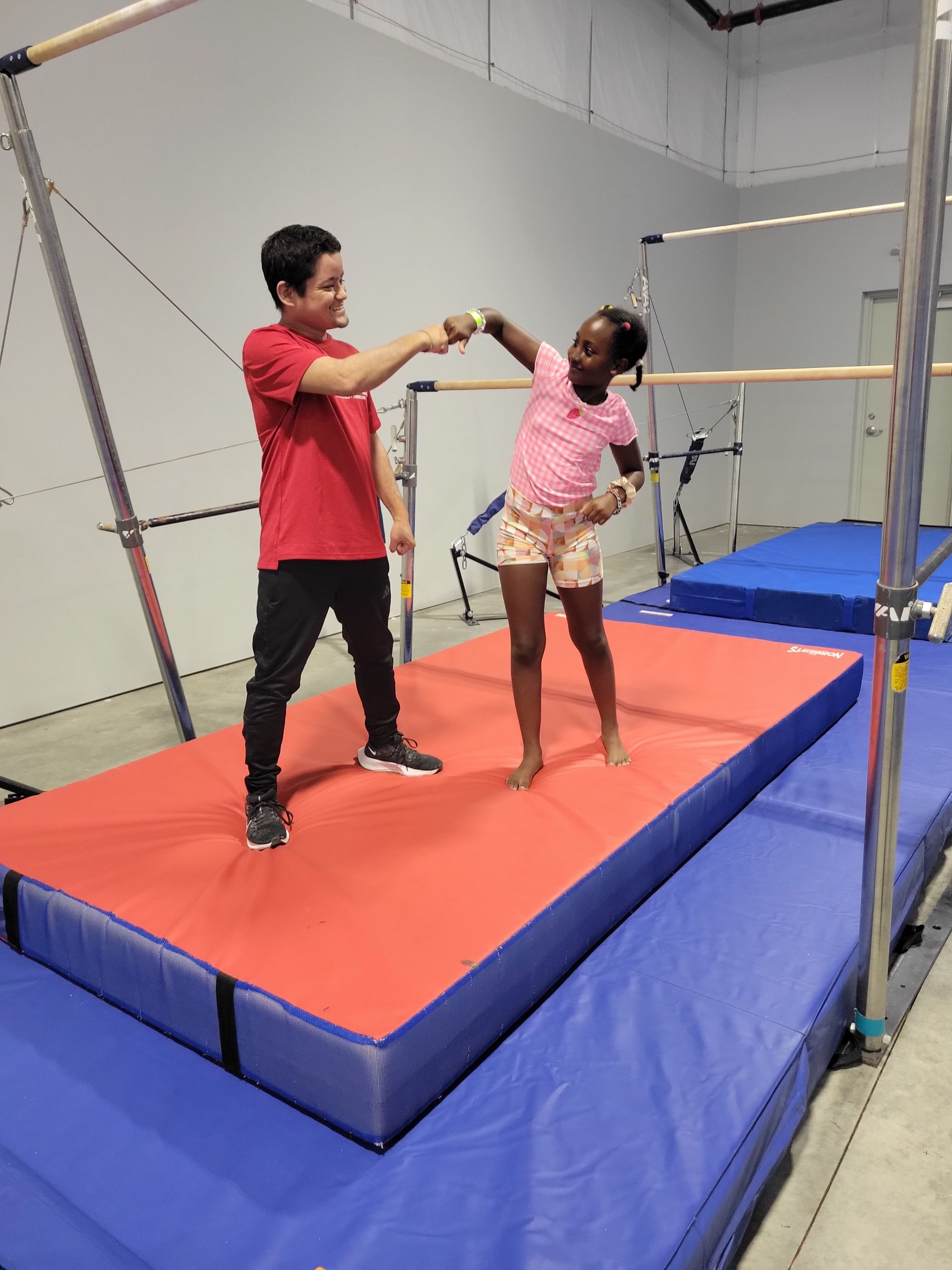 Gymnastics coach assisting a child on the uneven bars. Child in pink leotard, coach in black shirt and blue leggings.