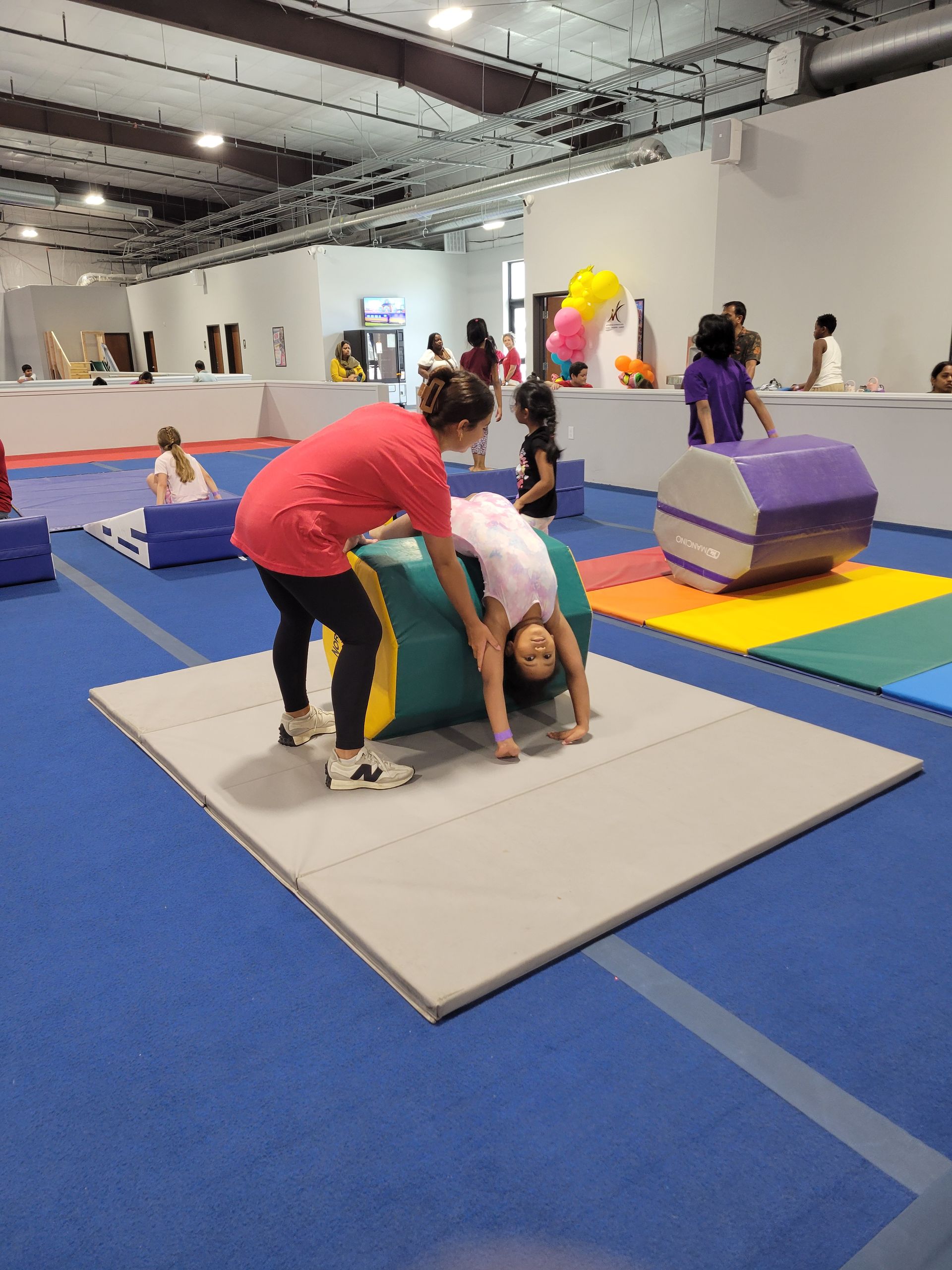 Gymnastics class: Instructor assisting a girl on a balance beam, other girls also on beams.