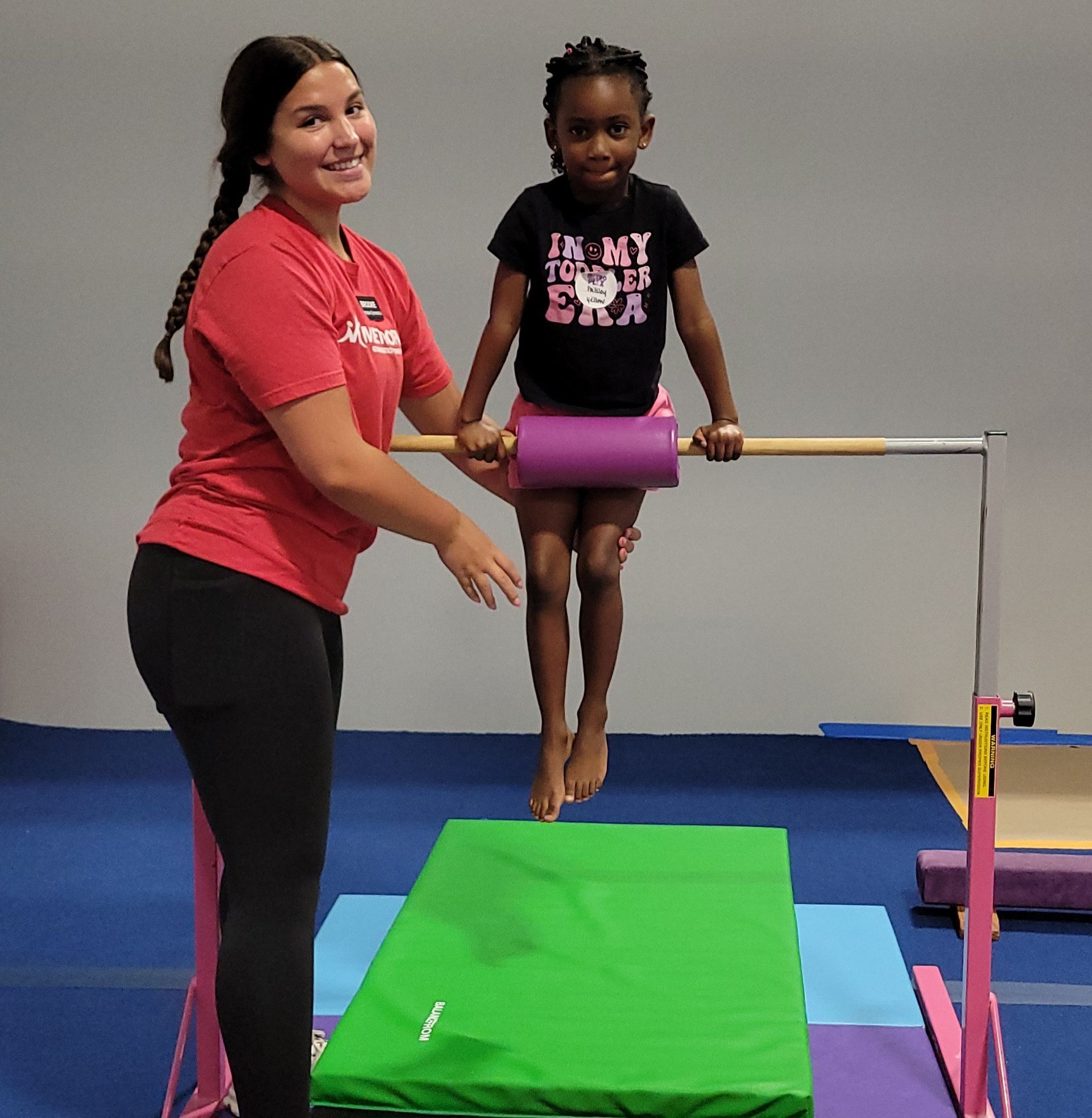 Woman assisting a child on a bar in a gymnastics gym; blue mats and equipment in the background.