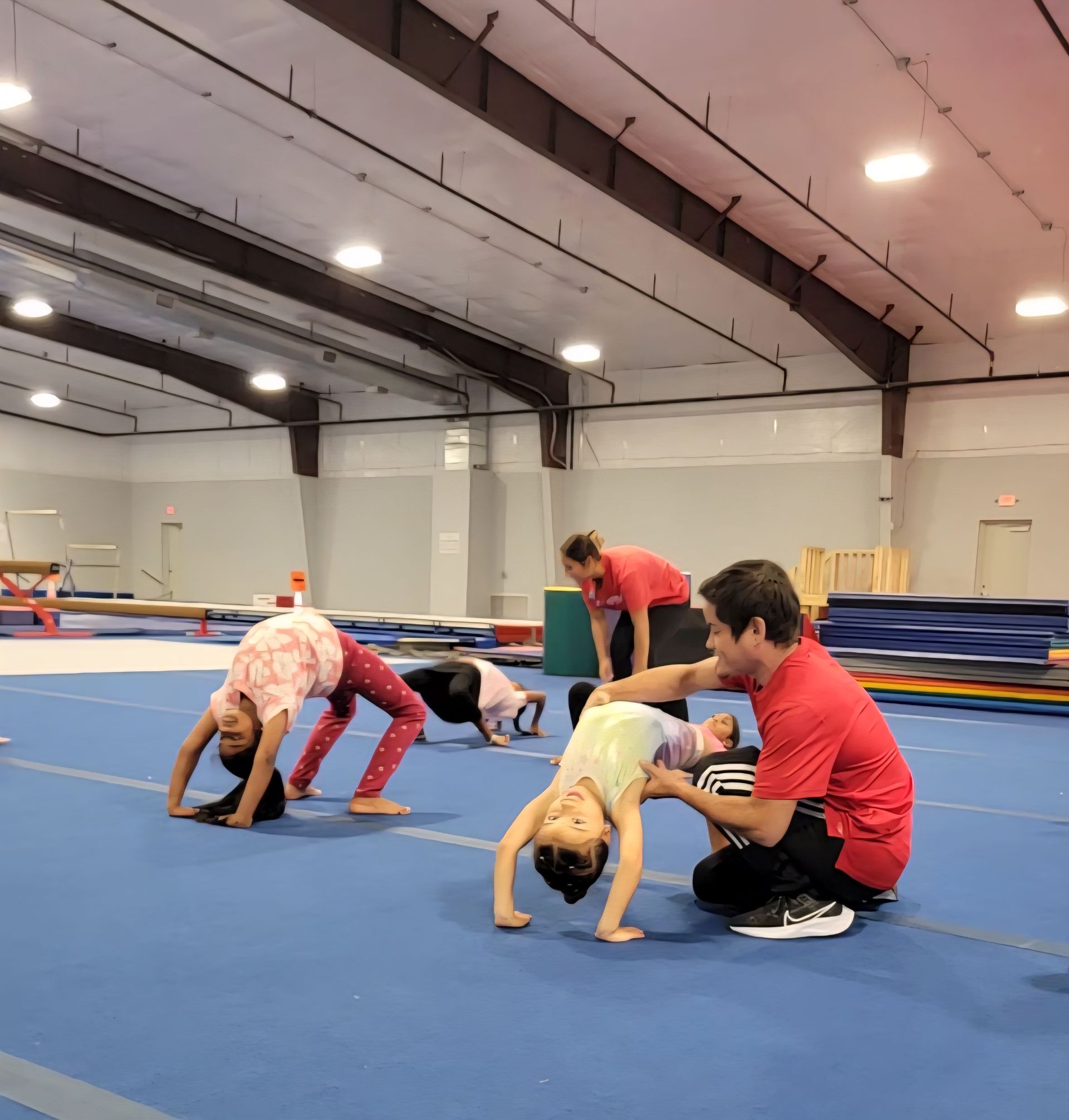 Two girls practicing gymnastics on balance beams with a coach in a gym.