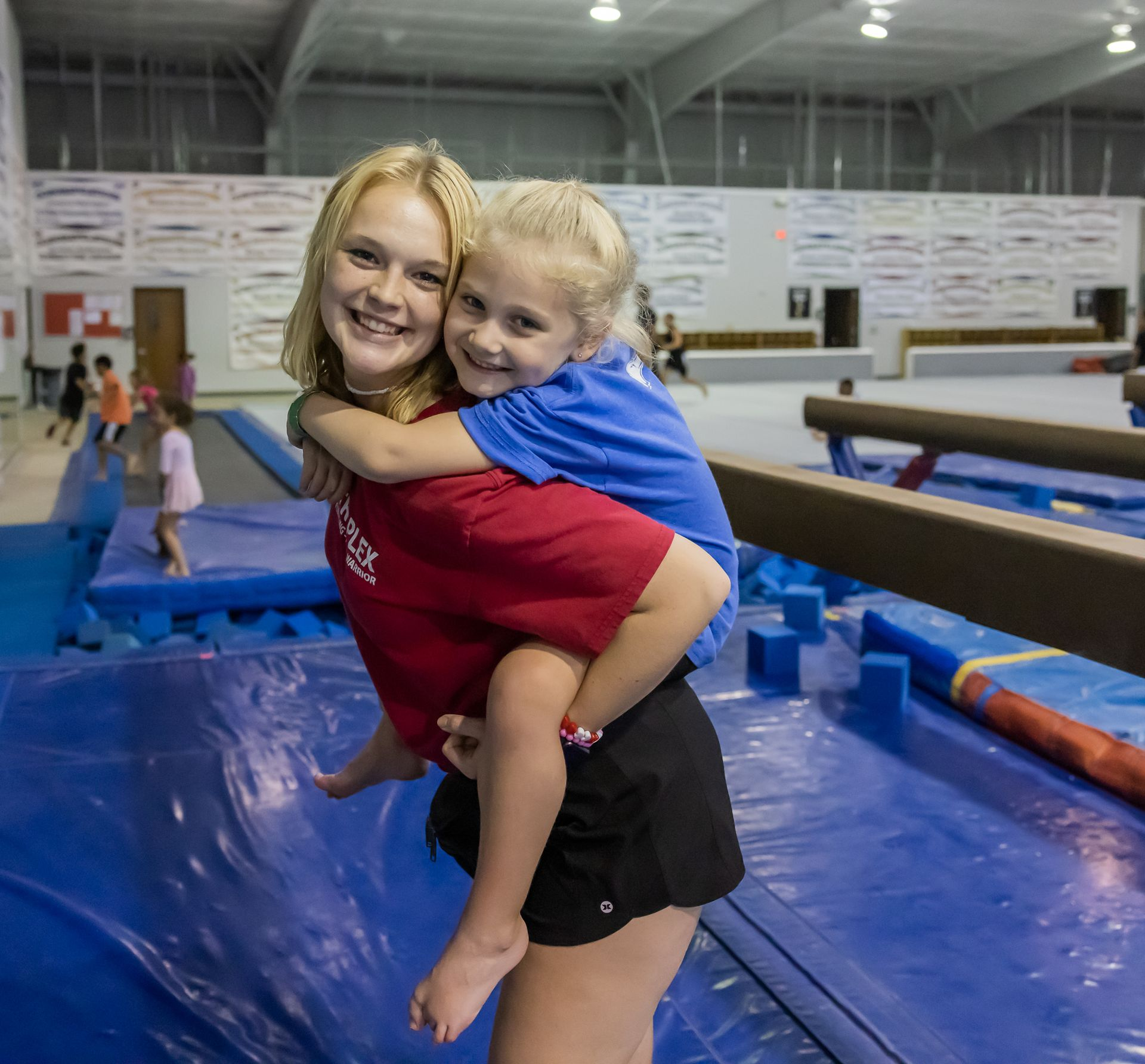 Two girls smiling, one giving the other a piggyback ride in a gymnastics gym, with mats and equipment.