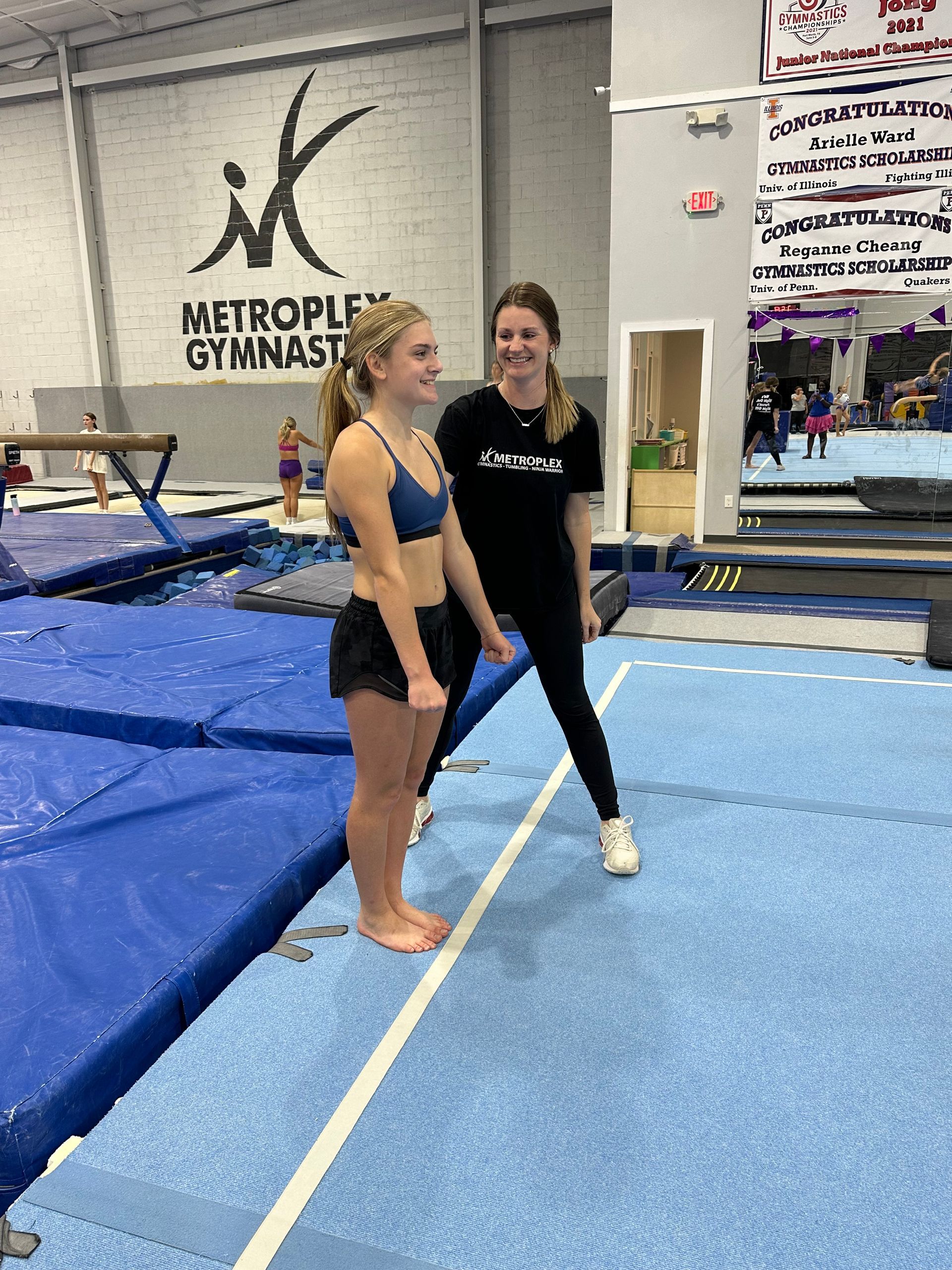 Gymnast smiling, standing with coach on blue mat in gymnasium. Black leotard.