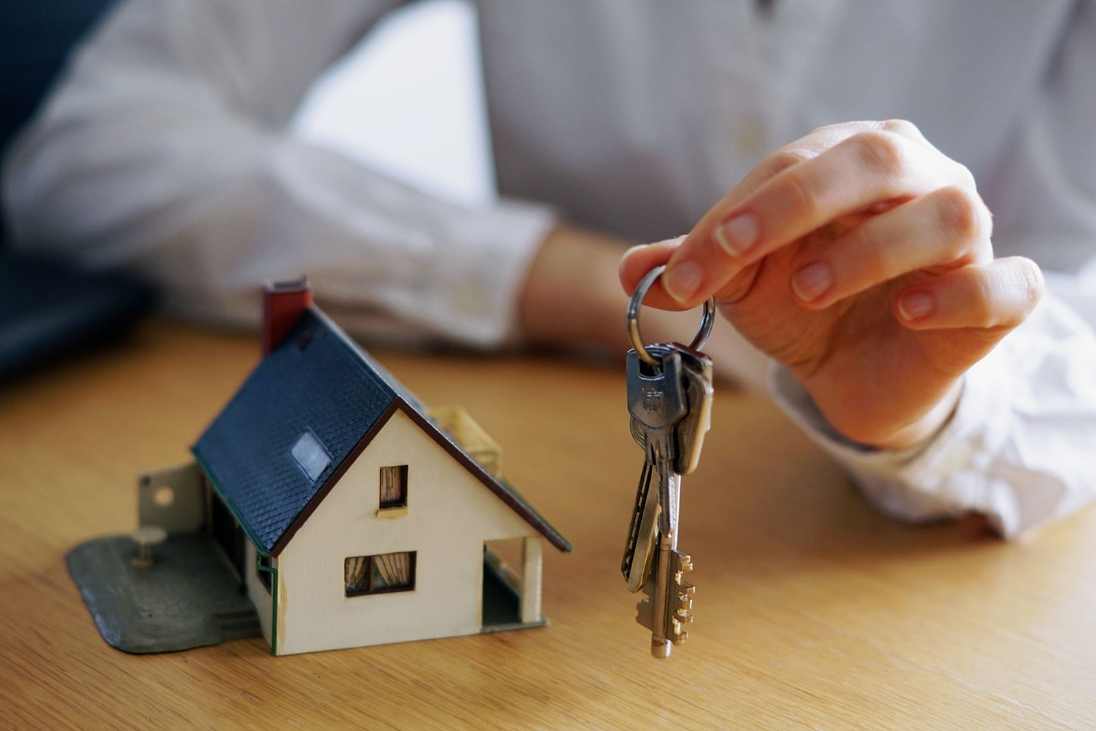 Persona sosteniendo llaves junto a un modelo de casa en miniatura sobre una mesa de madera.