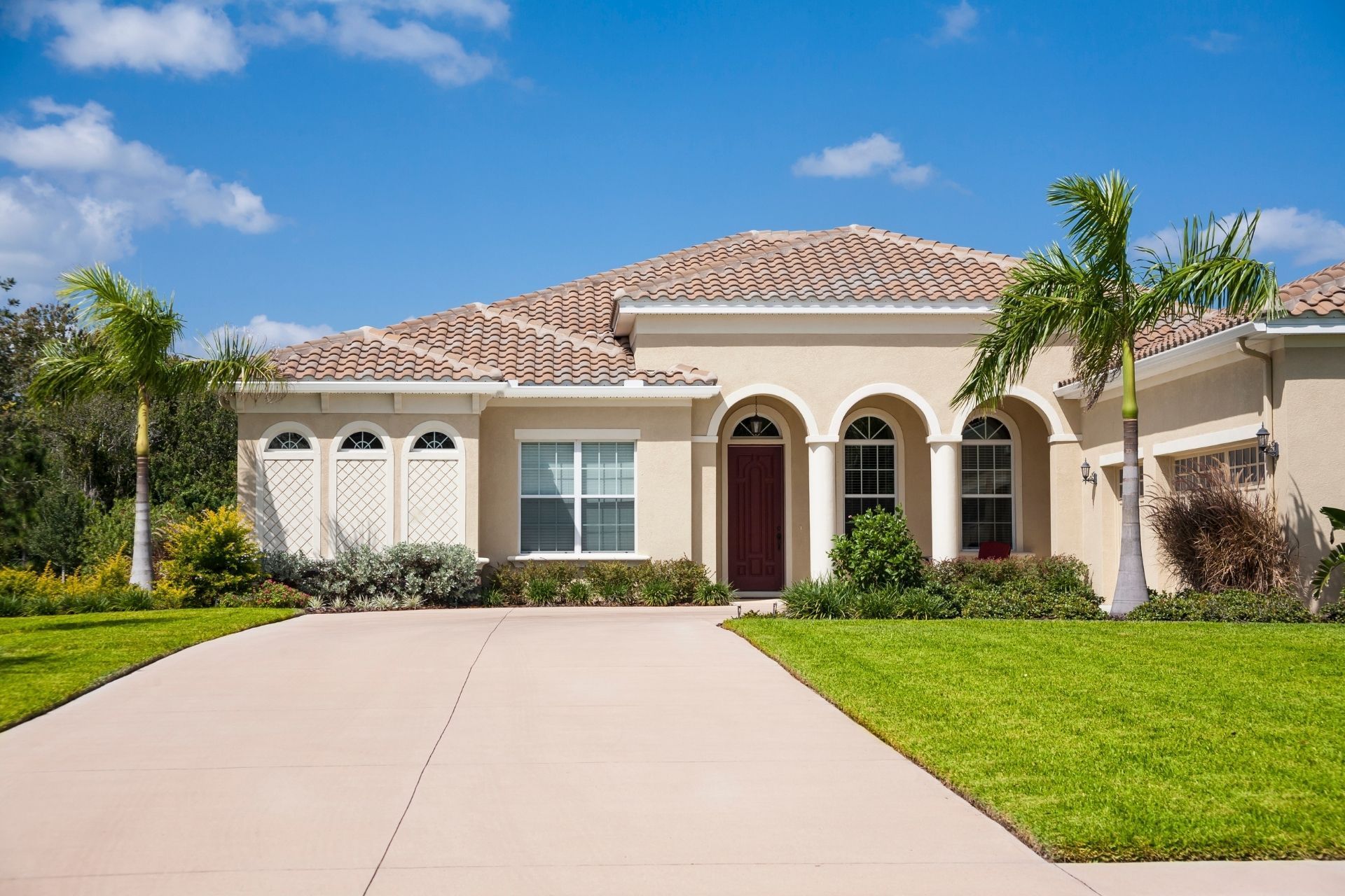Single-story house with a tiled roof, arched entry, and a wide driveway under a blue sky.