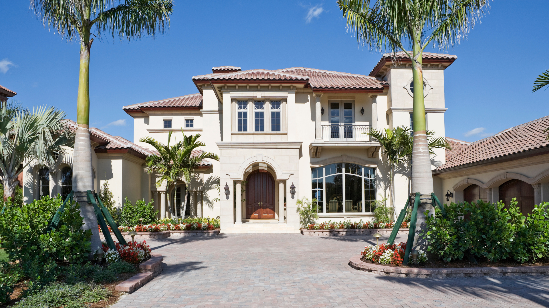 Luxury Mediterranean-style house with a tiled roof, palm trees, and a curved driveway under a clear blue sky