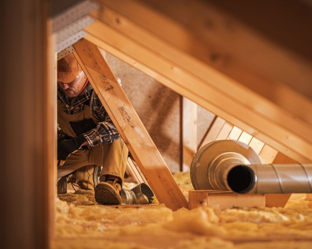 Worker crouching in an attic beside wooden rafters and HVAC ducting, with insulation covering the floor
