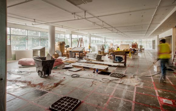 Construction site: room with debris, tools, and a worker sweeping. Large windows, concrete floor, and white walls.