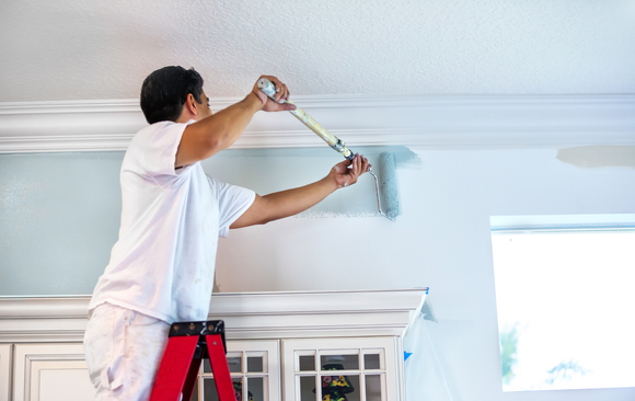 Man on a red ladder painting a light blue wall in a room, using a roller.