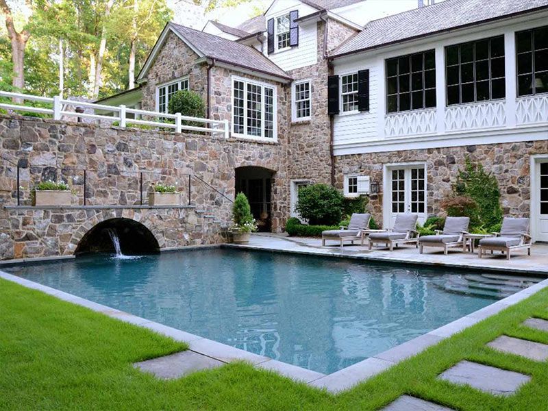Stone house with pool; arched bridge with waterfall, green lawn, and lounge chairs.