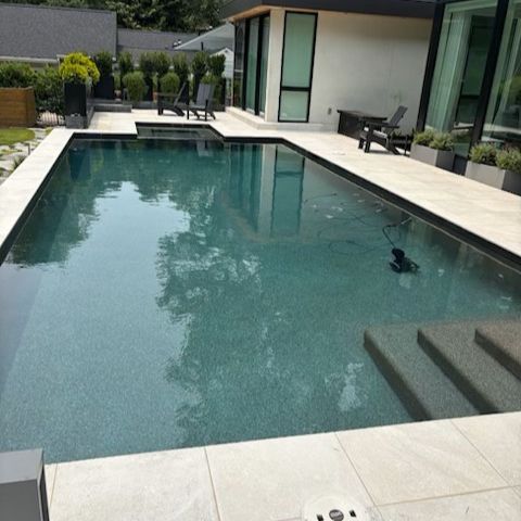 Rectangular pool with dark water, bordered by light-colored tiles. Modern house in background, with outdoor seating.