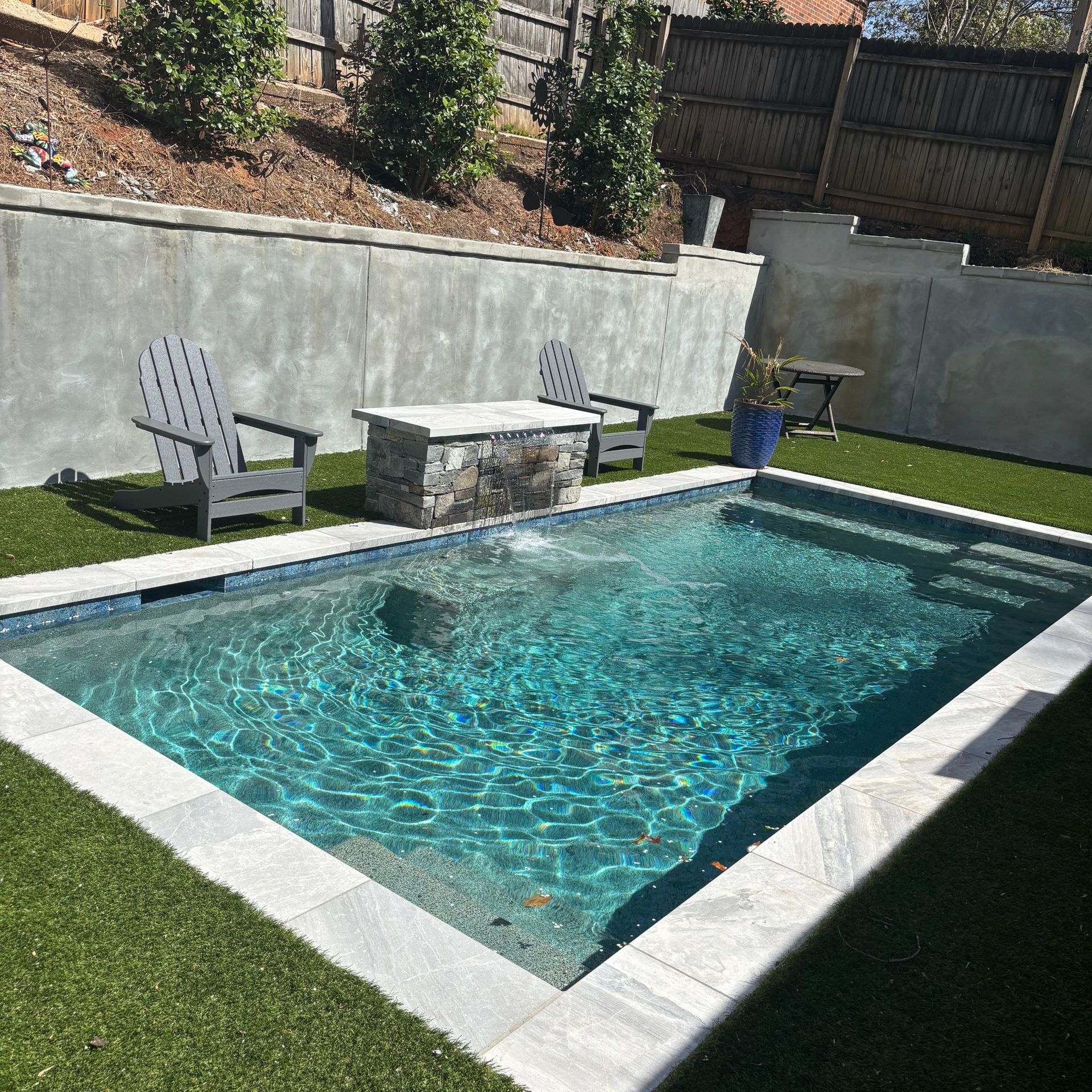 Small backyard pool with waterfall, two chairs, and artificial turf.