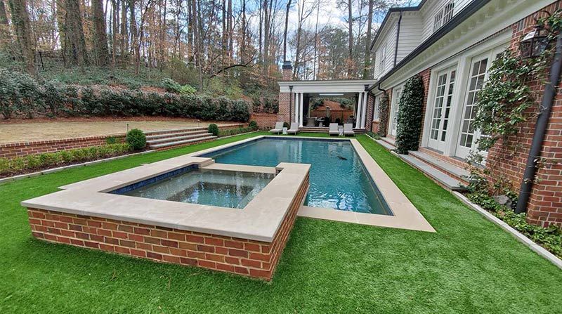 A pool and spa area with a brick border, green grass, and a home in the background.