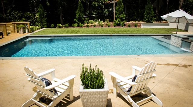 Two white Adirondack chairs face a rectangular pool, with a hedge in the foreground and trees in the background.