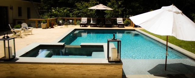 Outdoor pool with a hot tub, lounge chairs, umbrellas, and trees.