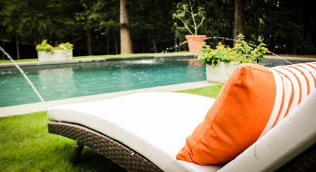 Lounge chair with orange pillow by a pool with fountains and potted plants.