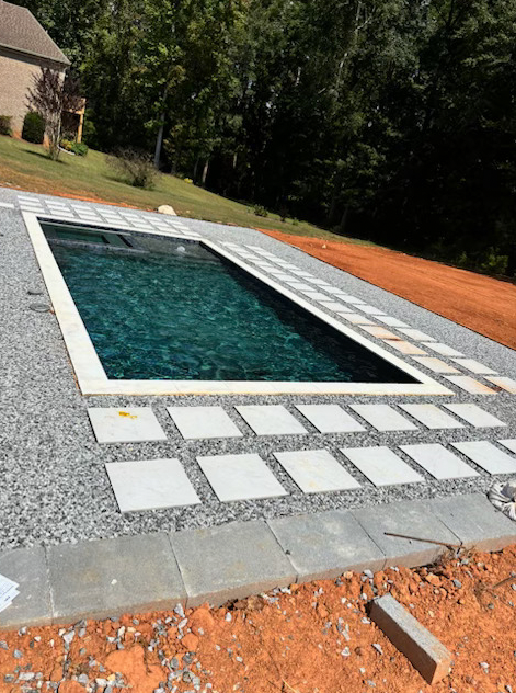 Rectangular pool with dark water, surrounded by gray gravel and square concrete tiles in a yard.