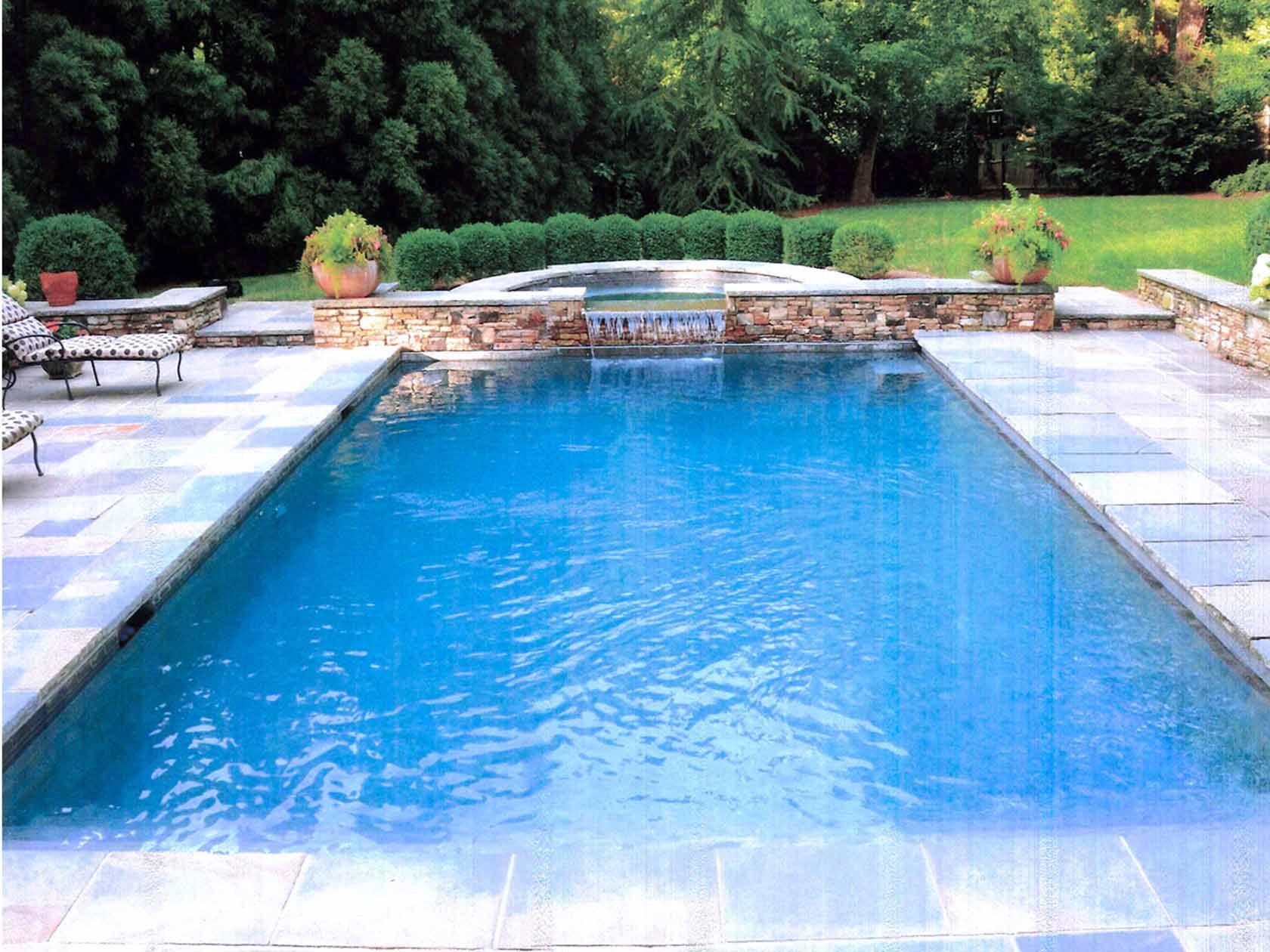 Blue rectangular swimming pool with stone patio, waterfall, and manicured lawn.