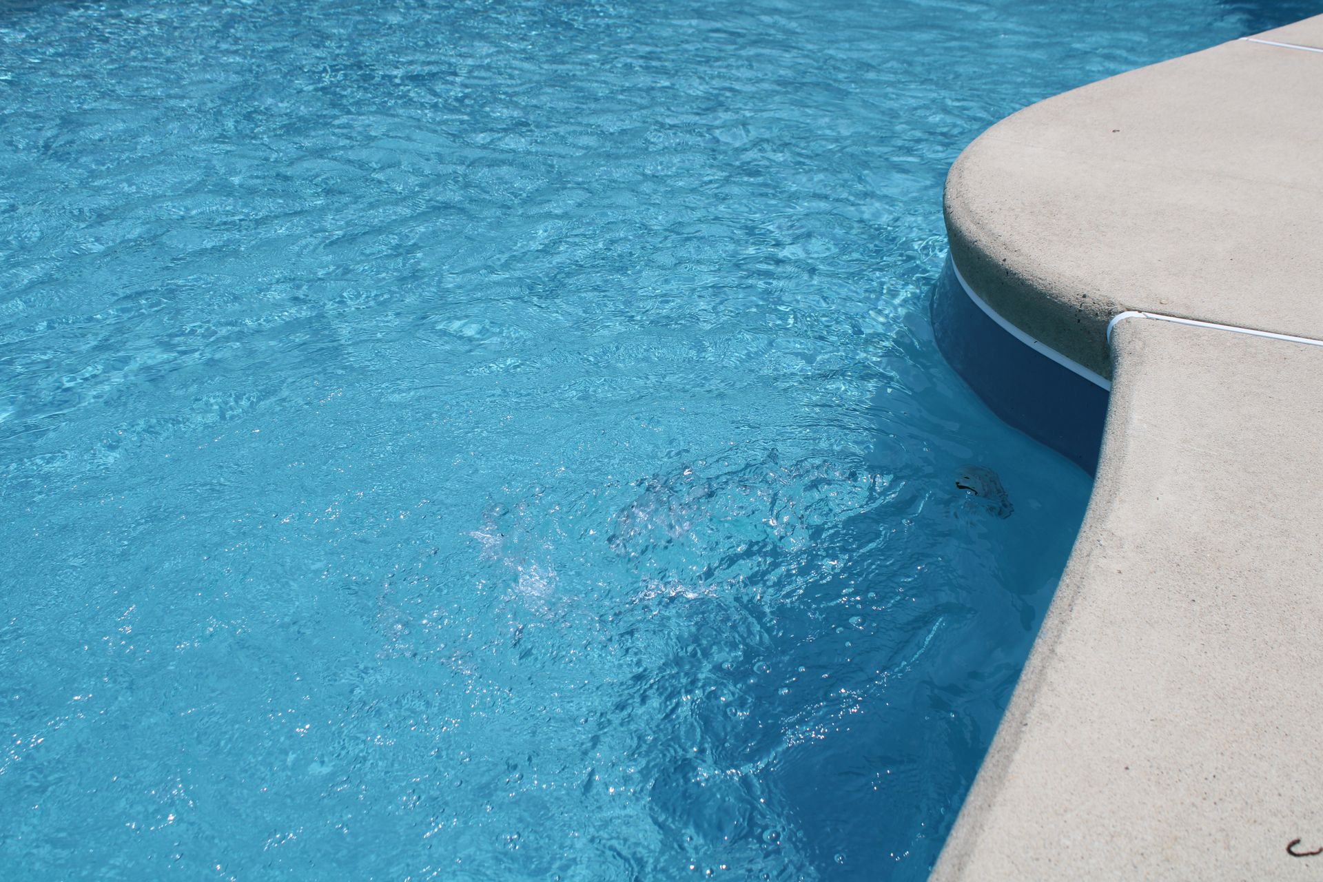 Blue pool water next to a concrete edge; the water sparkles in the sunlight.