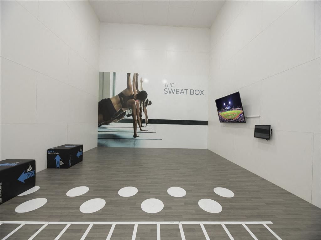 Interior fitness studio with a mural reading 'The Sweat Box', wooden floor, and wall-mounted TV at River Oaks.