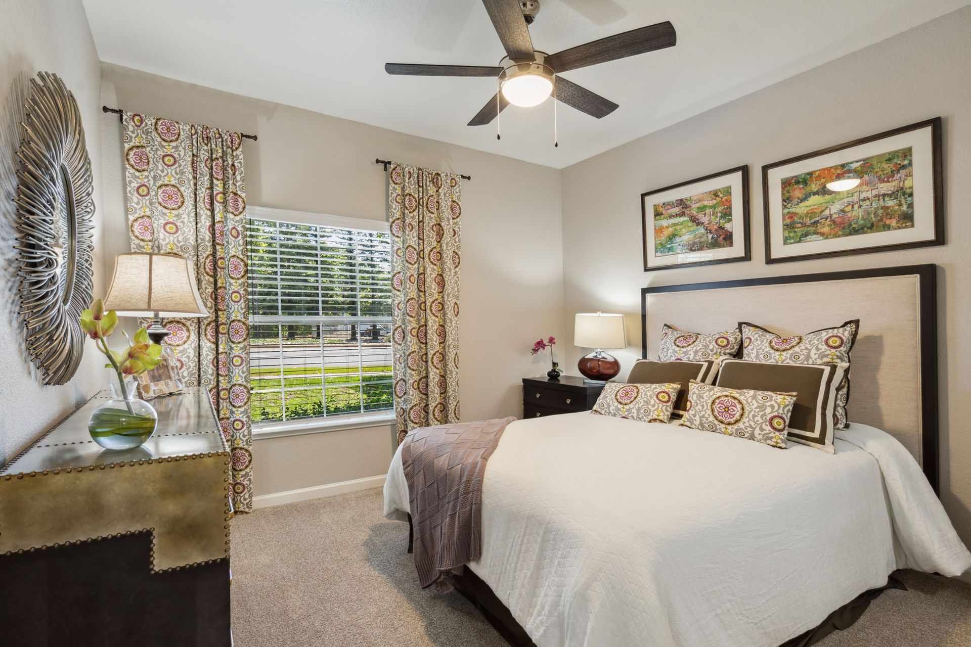 Bedroom with a bed, nightstand, and window with patterned curtains; neutral walls and carpet at River Oaks.
