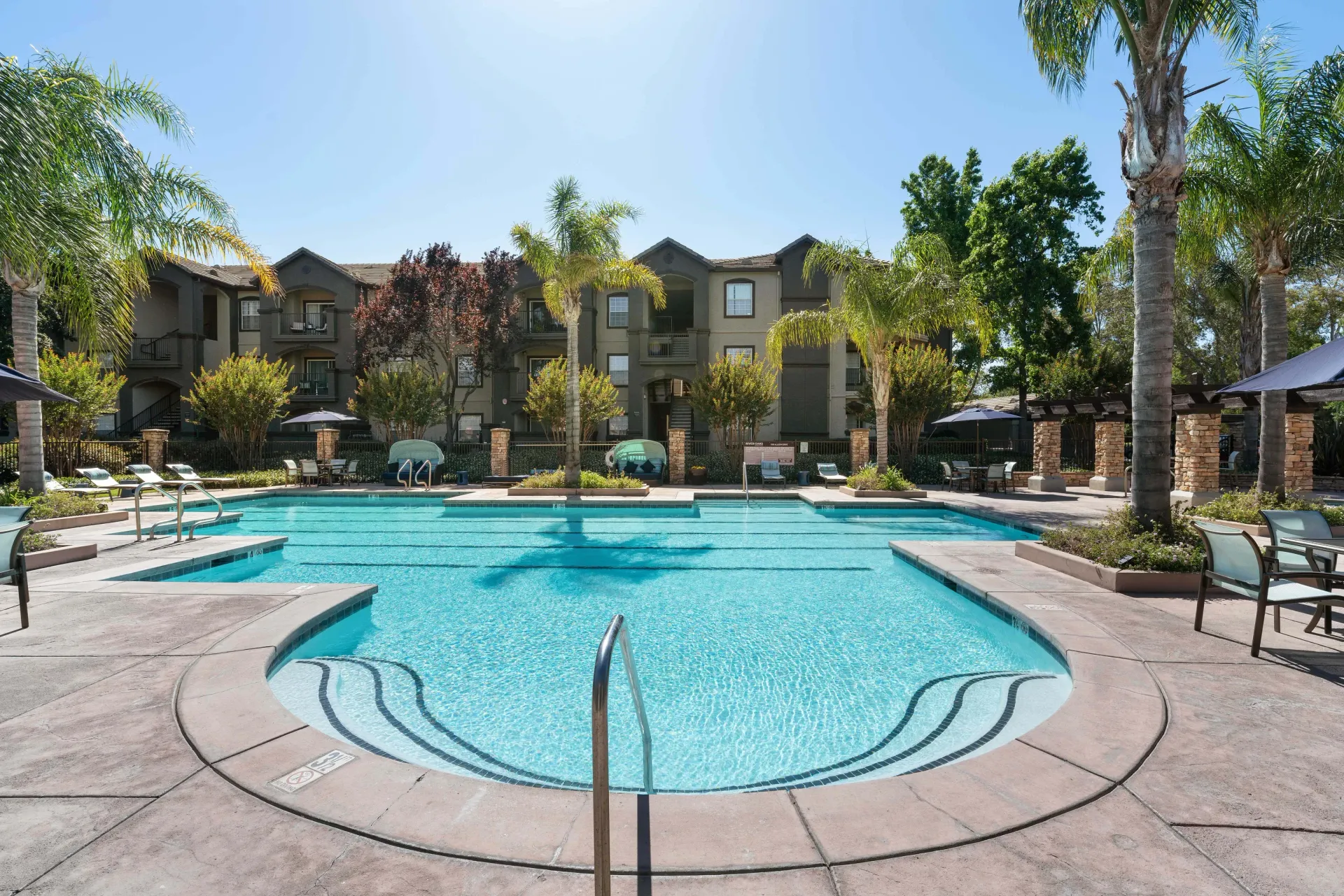 Swimming pool in front of apartment buildings on a sunny day, with palm trees and lounge chairs at River Oaks.