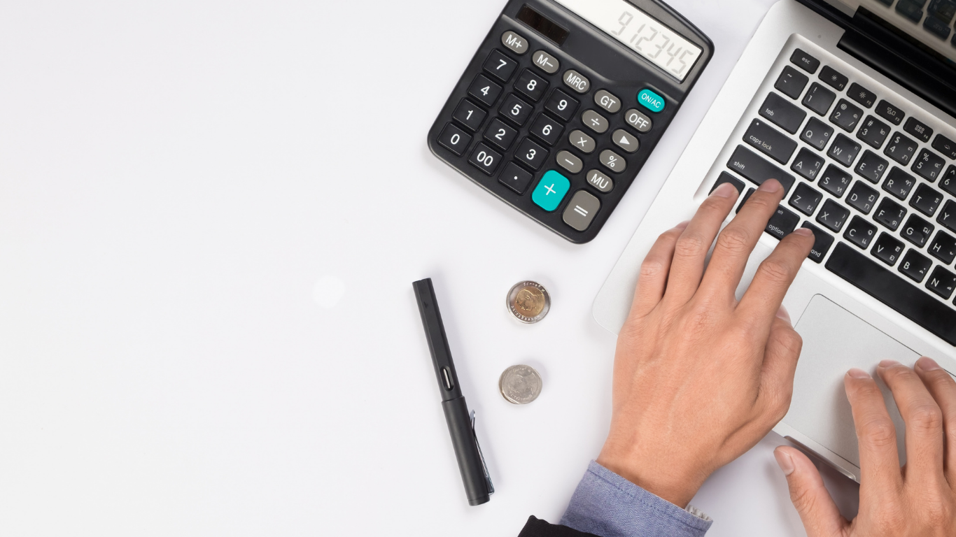 Hands typing on laptop beside calculator, pen, and coins on a white desk.