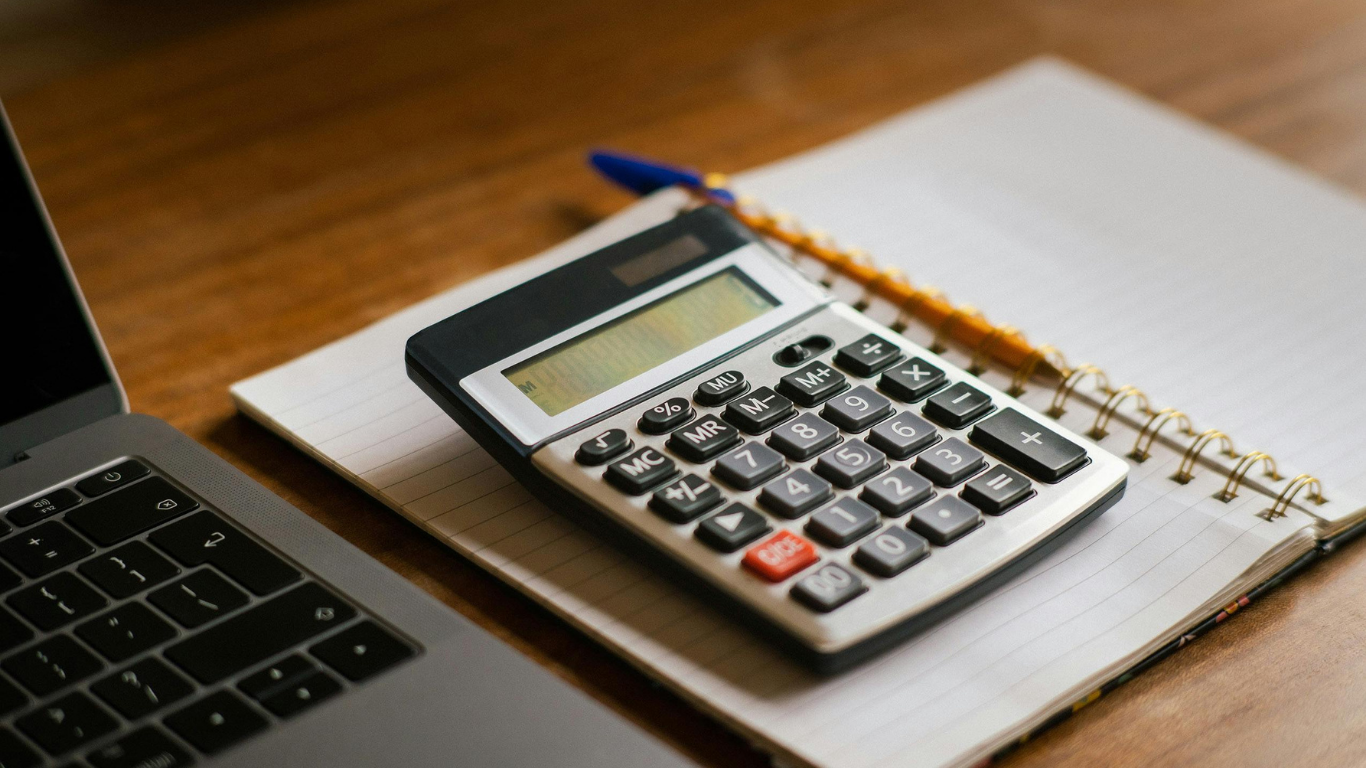 Calculator on a spiral notebook beside a laptop on a wooden desk