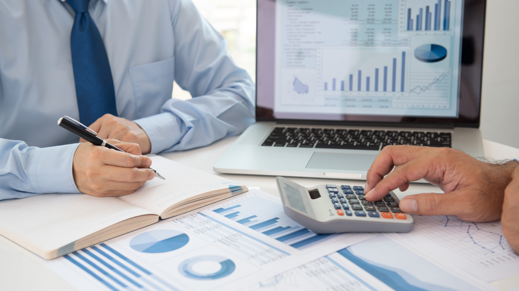 Hands using a calculator and pen over financial charts and notes on a desk