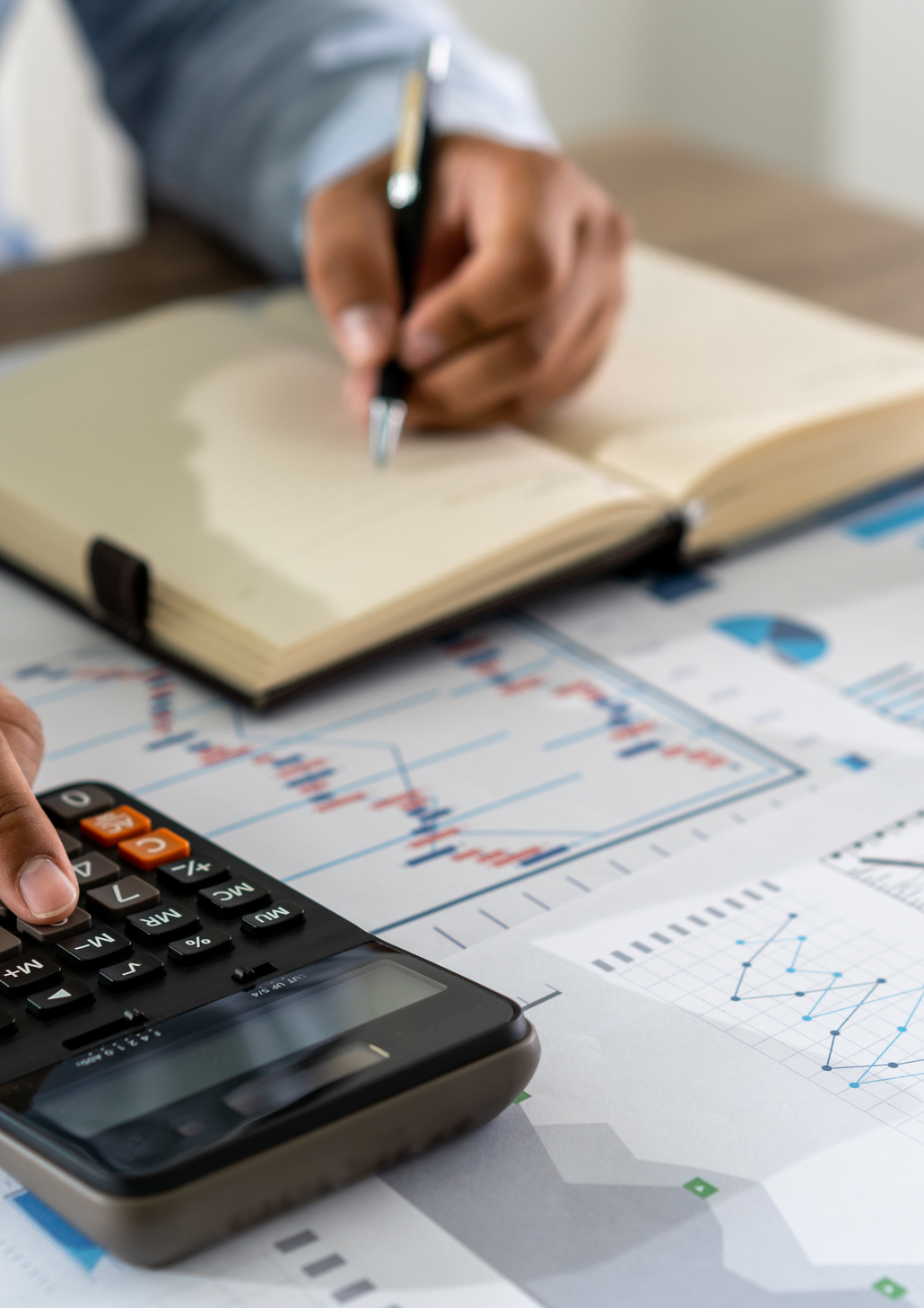 Hand writing notes beside a calculator and financial charts on a desk