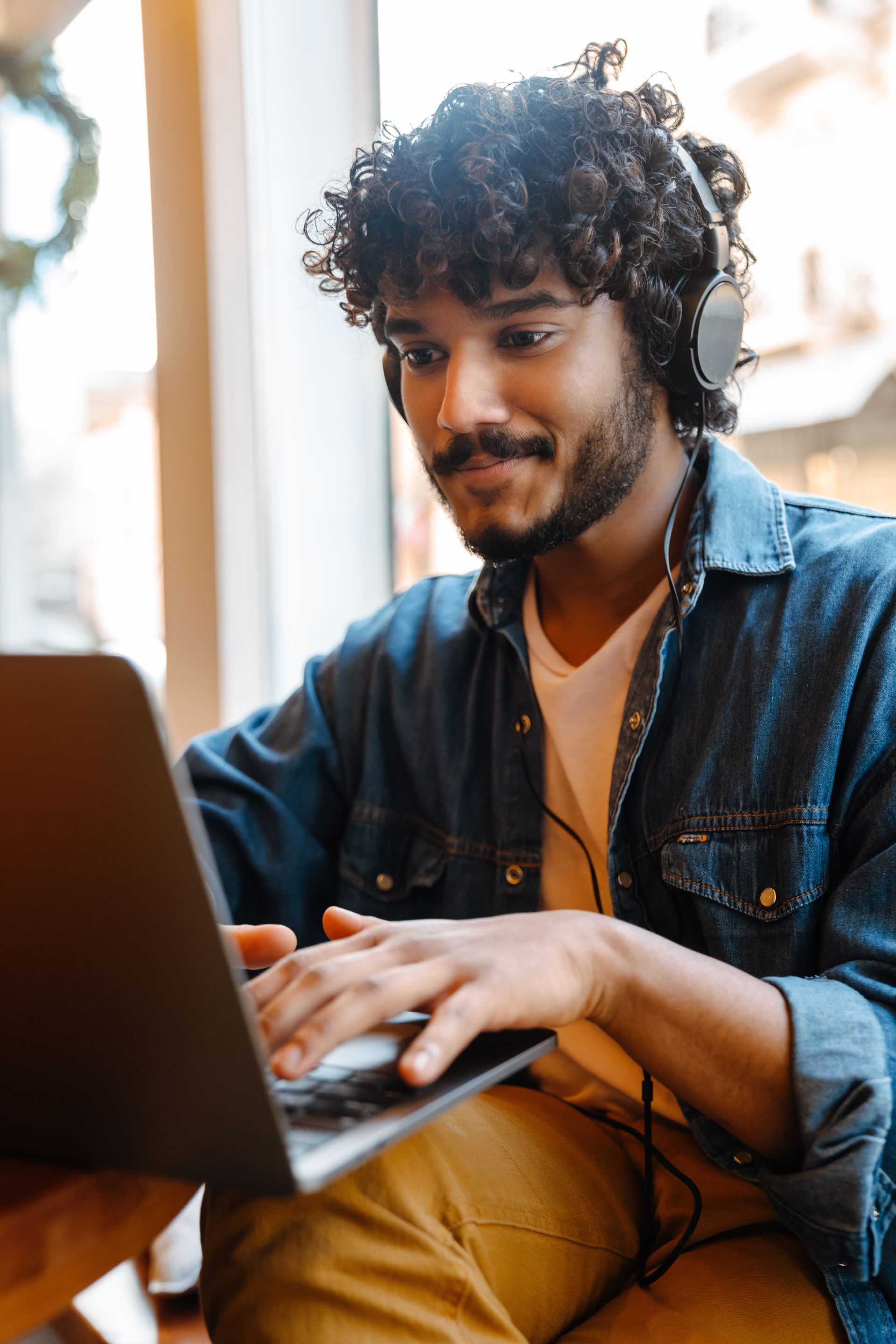Person typing on a laptop, wearing headphones and a denim shirt in a bright indoor setting
