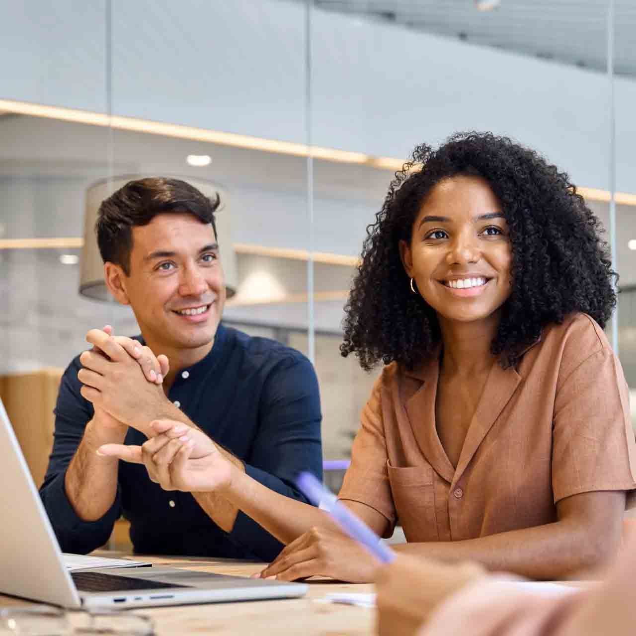 Deux collègues sourient dans un bureau moderne, assis à une table avec un ordinateur portable et des notes.