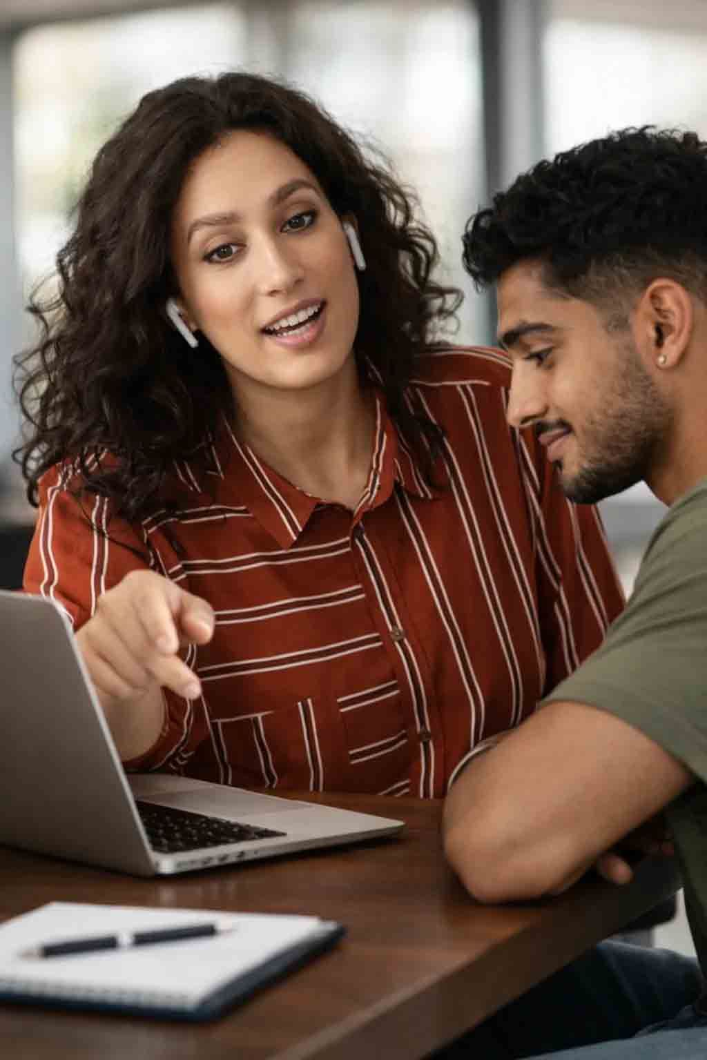 Two people discussing a laptop at a table, with a notebook in the foreground.