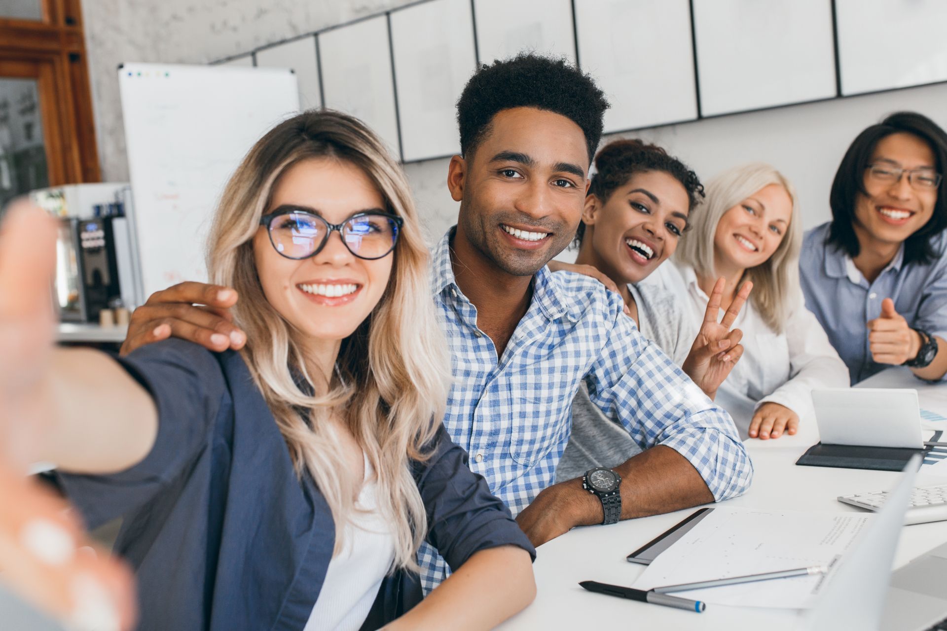 Five coworkers smiling at a desk, taking a selfie in a bright office with laptops and notebooks