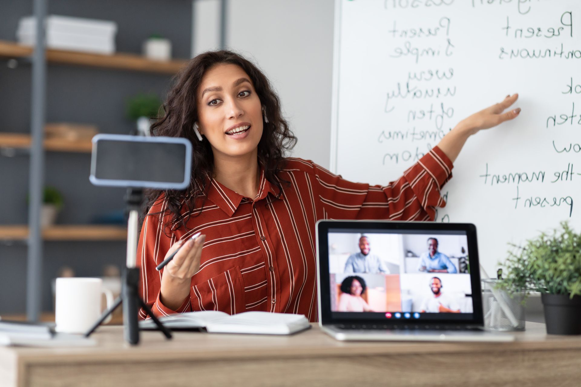 Une femme, assise à un bureau, donne un cours en ligne et désigne un tableau blanc, une visioconférence étant installée devant elle.