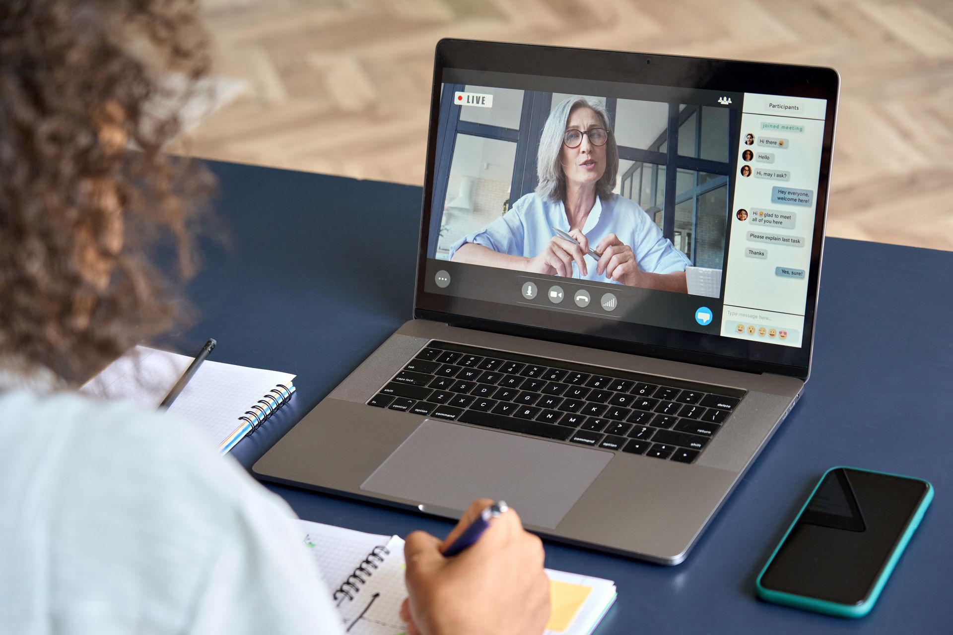 Person in a video call on a laptop, writing notes beside a smartphone on a desk.