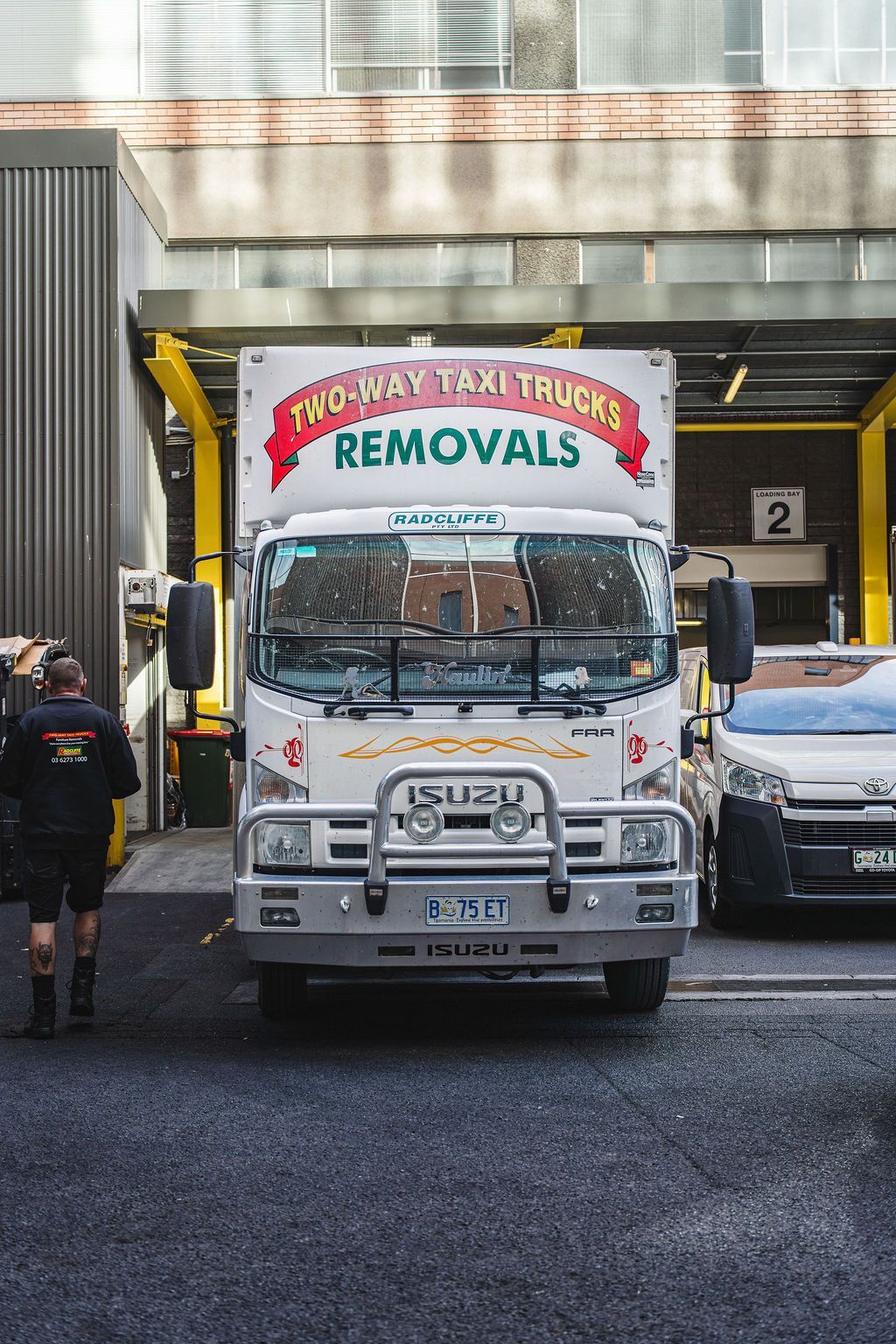 White removals truck parked under an overhang, with a person walking beside it and a car behind.