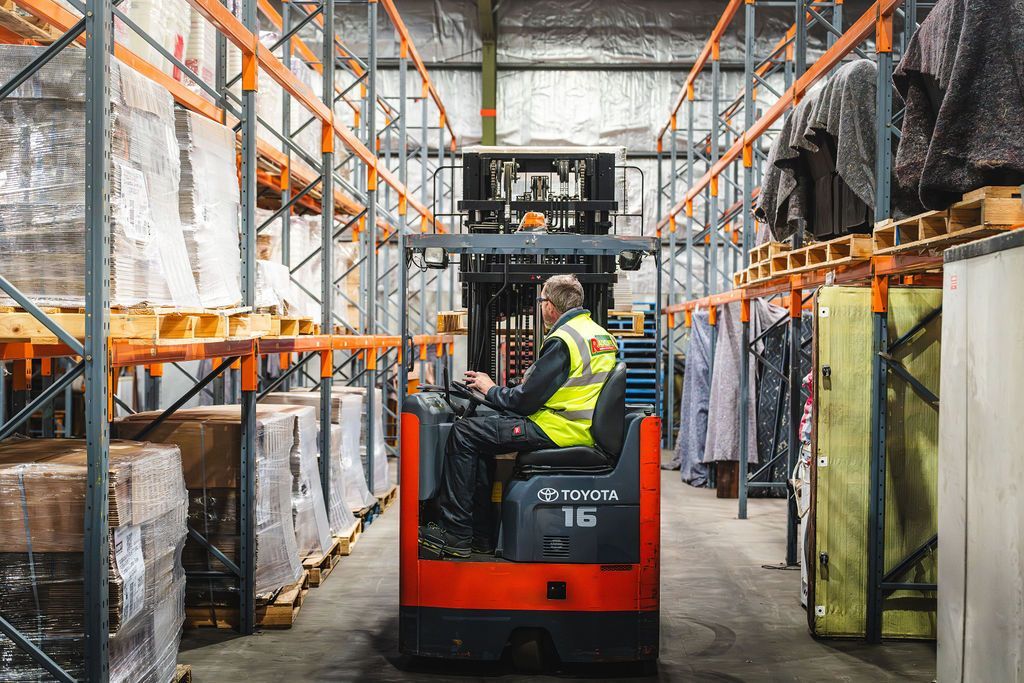 Worker driving a red forklift in a warehouse aisle lined with stacked goods and pallets