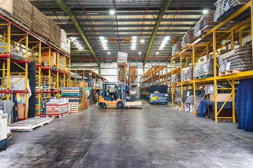 Large warehouse aisle with stacked goods, forklifts, and tall yellow shelving under bright industrial lights