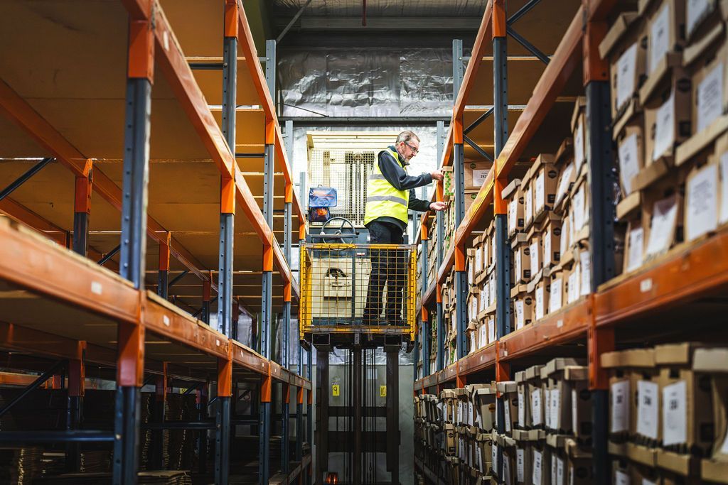 Forklift lifting a worker in a warehouse aisle between tall orange and blue storage racks