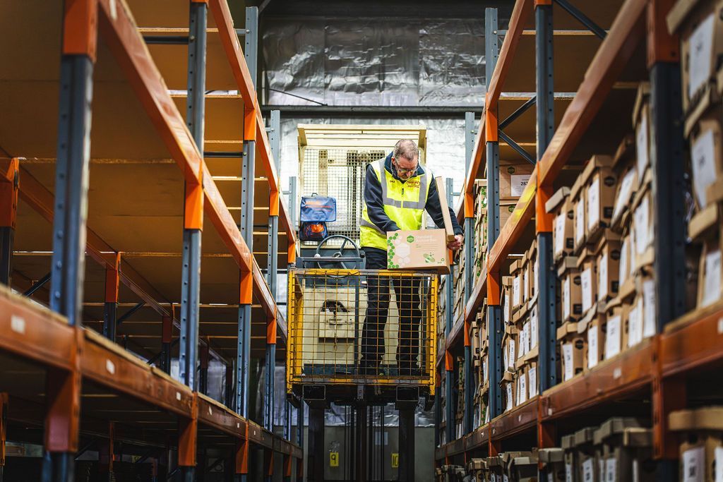 Workers on a lift inspecting warehouse shelves with stacked boxes on both sides