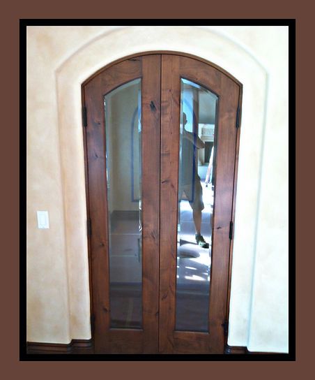 Double wooden doors with glass panes, arched top, tan walls, person reflected in glass.