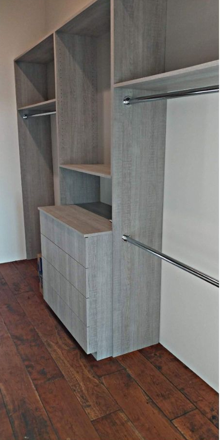 A light gray, wood-grain closet organizer with shelves, hanging rods, and a drawer unit on a dark wooden floor.