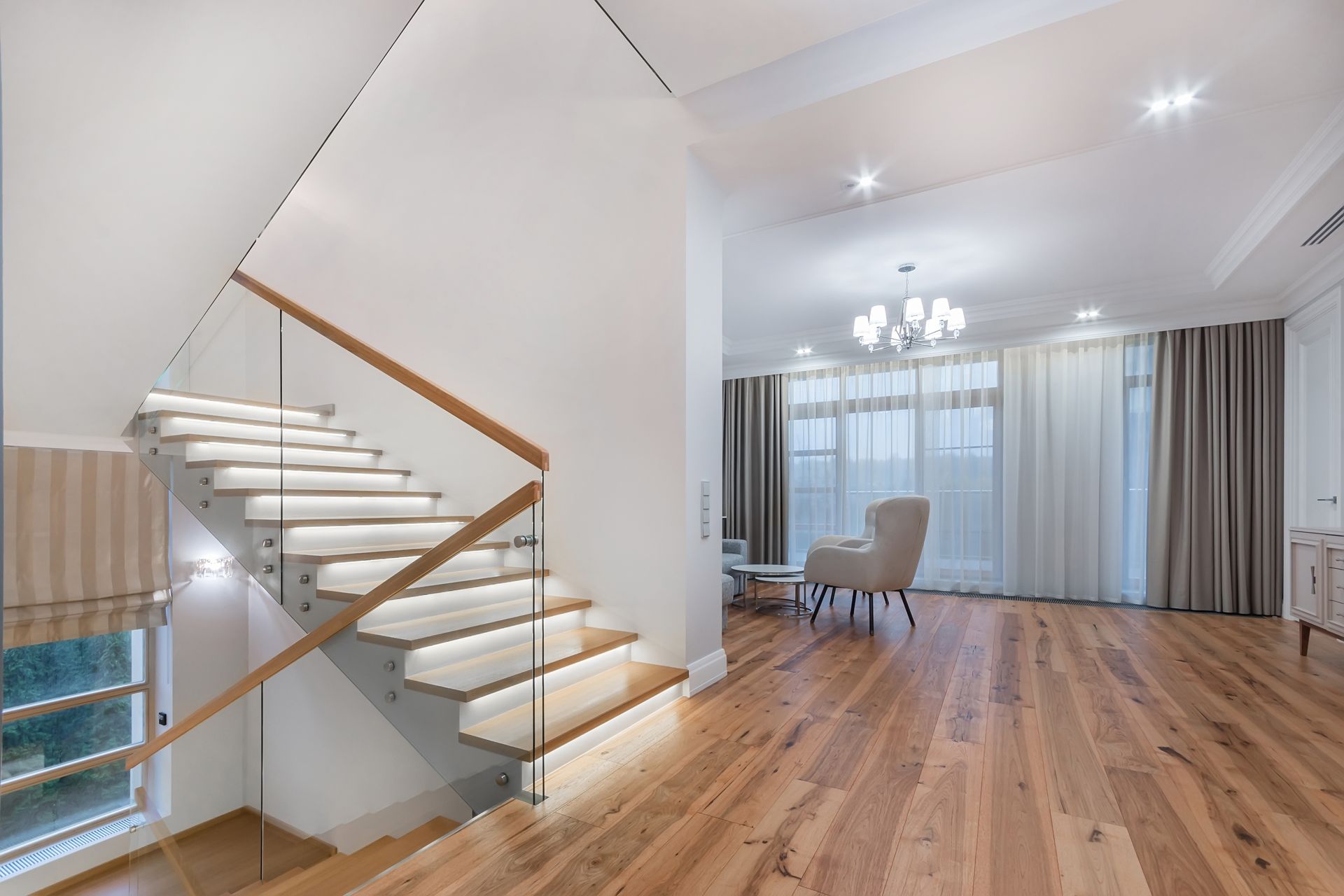 Modern living room with hardwood floors, floating staircase, chandelier, and large windows with curtains