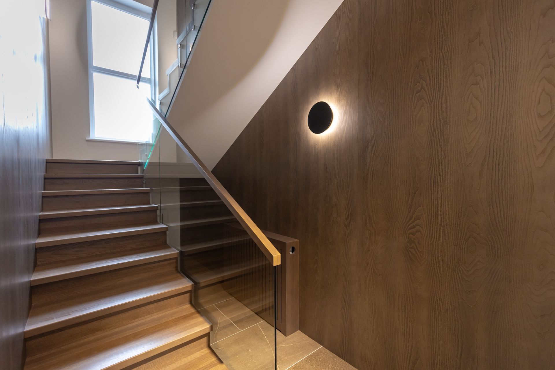 Modern stairwell with wooden steps, glass railing, and round wall light on a brown wall
