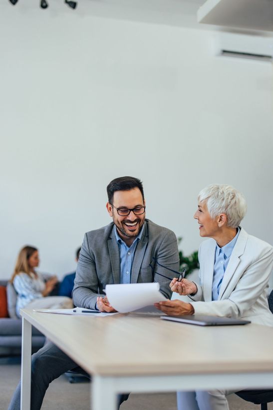 Two coworkers smiling and talking over papers at a conference table in a bright office