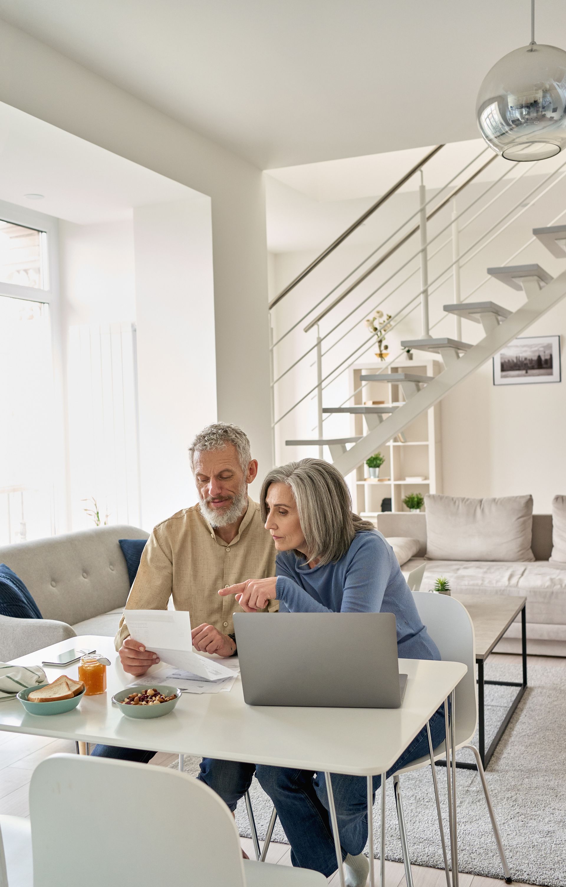 Two people working on a laptop at a table in a bright living room with a staircase behind them